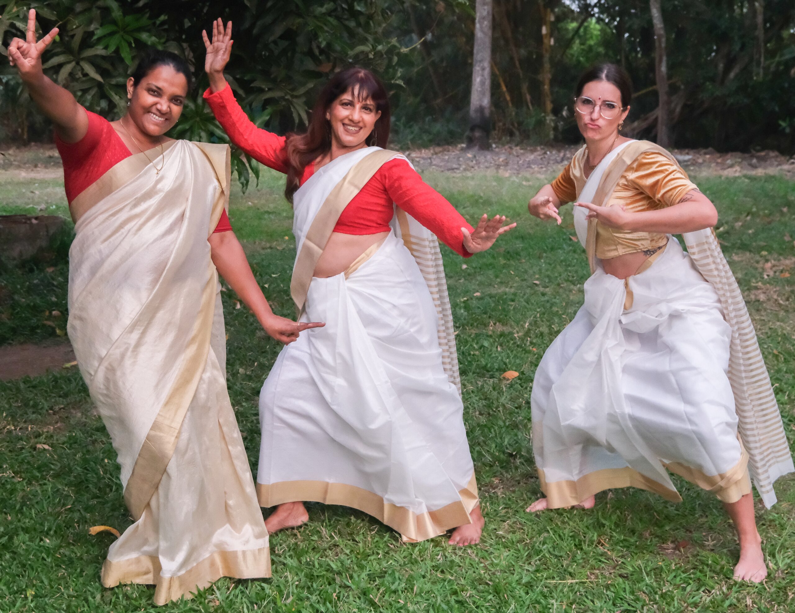 Women dressed in traditional gold-trimmed off-white saris dancing in a circle around a brass lamp during a somatic Thiruvathirakali sisterhood dance at Sangeeta Pillai's Devi Sisterhood retreat